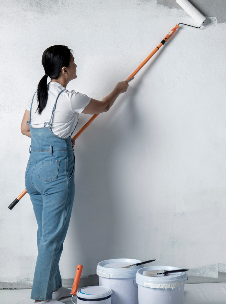 Female painter applying white paint on a new wall using roller and extension pole during interior finishing work.